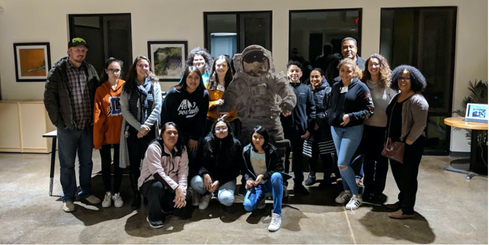 Fifteen people pose around a cardboard cutout of an astronaut in moon landing suit. Three kneel in front, six stand to the left, and six stand to the right. They are a mix of adults and youths, likely high school-aged.