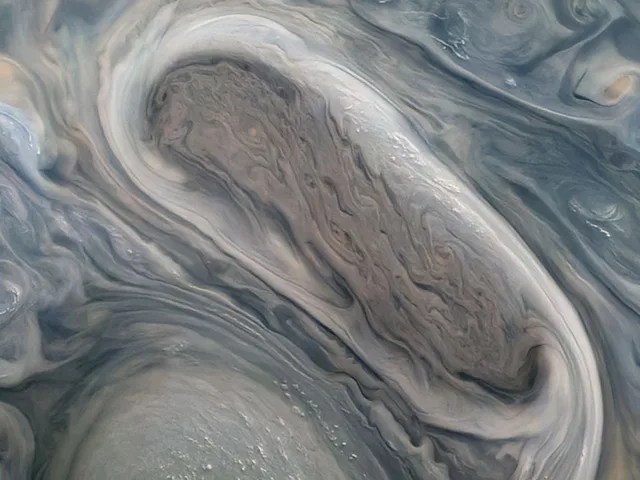 Zoomed in view of Jupiter's surface. An oblong grey oval is shown between swirling clouds.
