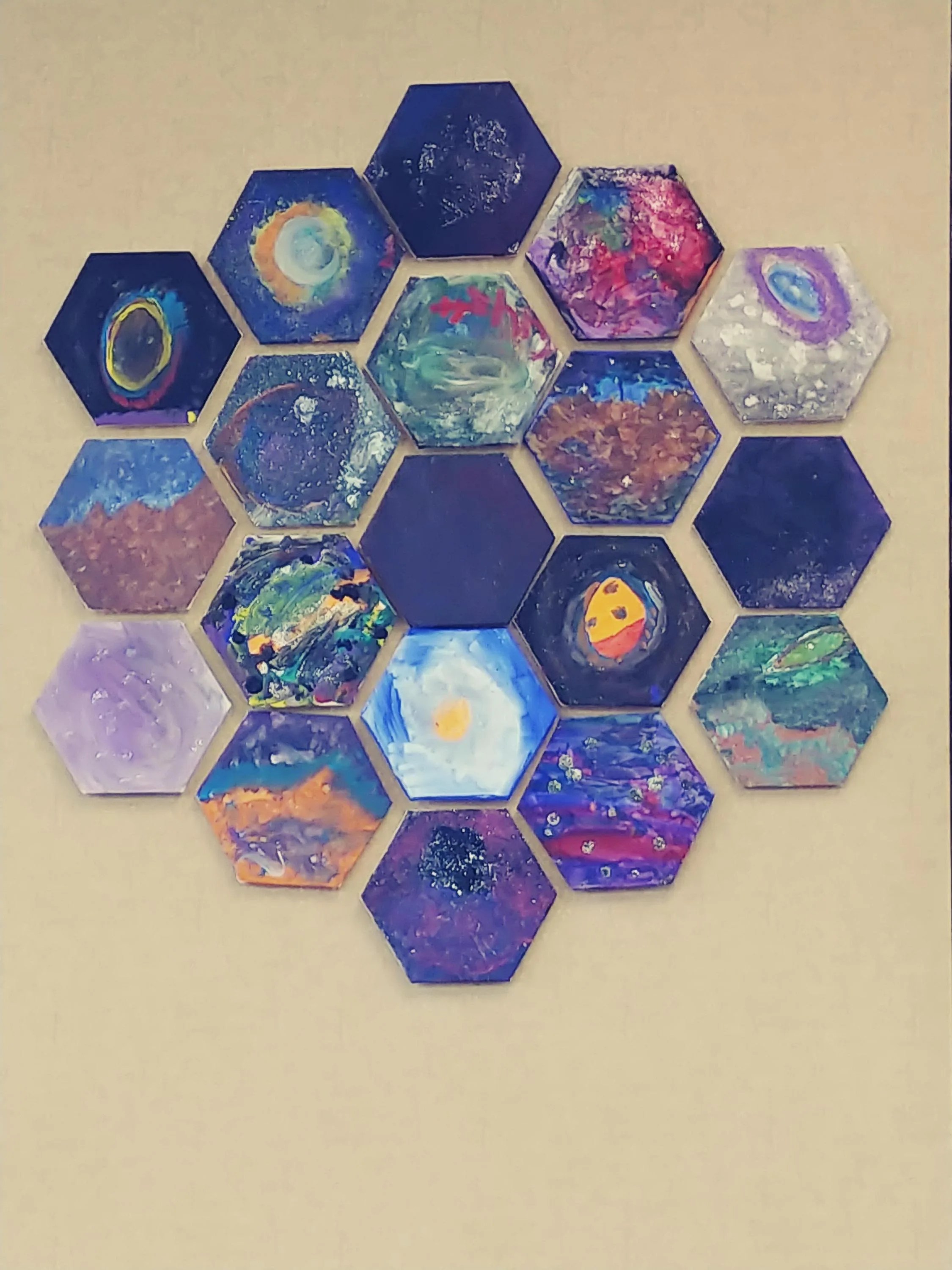 19 small canvas hexagons are arranged in one large hexagon similar in share to the James Webb Space Telescope Mirror handing on a wall. Each individual hexagon is different with swirls of blue, white, purple, red, gray depict galaxies, nebulas, and other space elements.