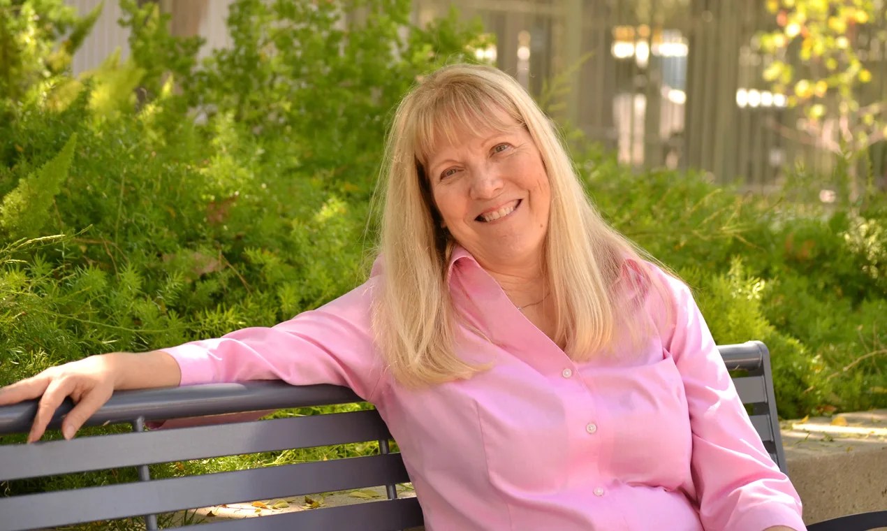 Linda is sitting with her arm outstretched along a park bench. She's wearing a bright pink shirt and the sun in shining through green leaves of surrounding trees.