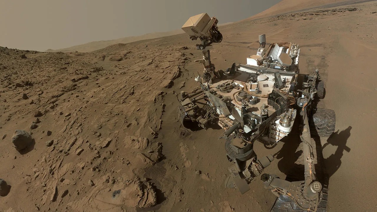 A rover covered in dust sits on a rocky, sandy surface on Mars.