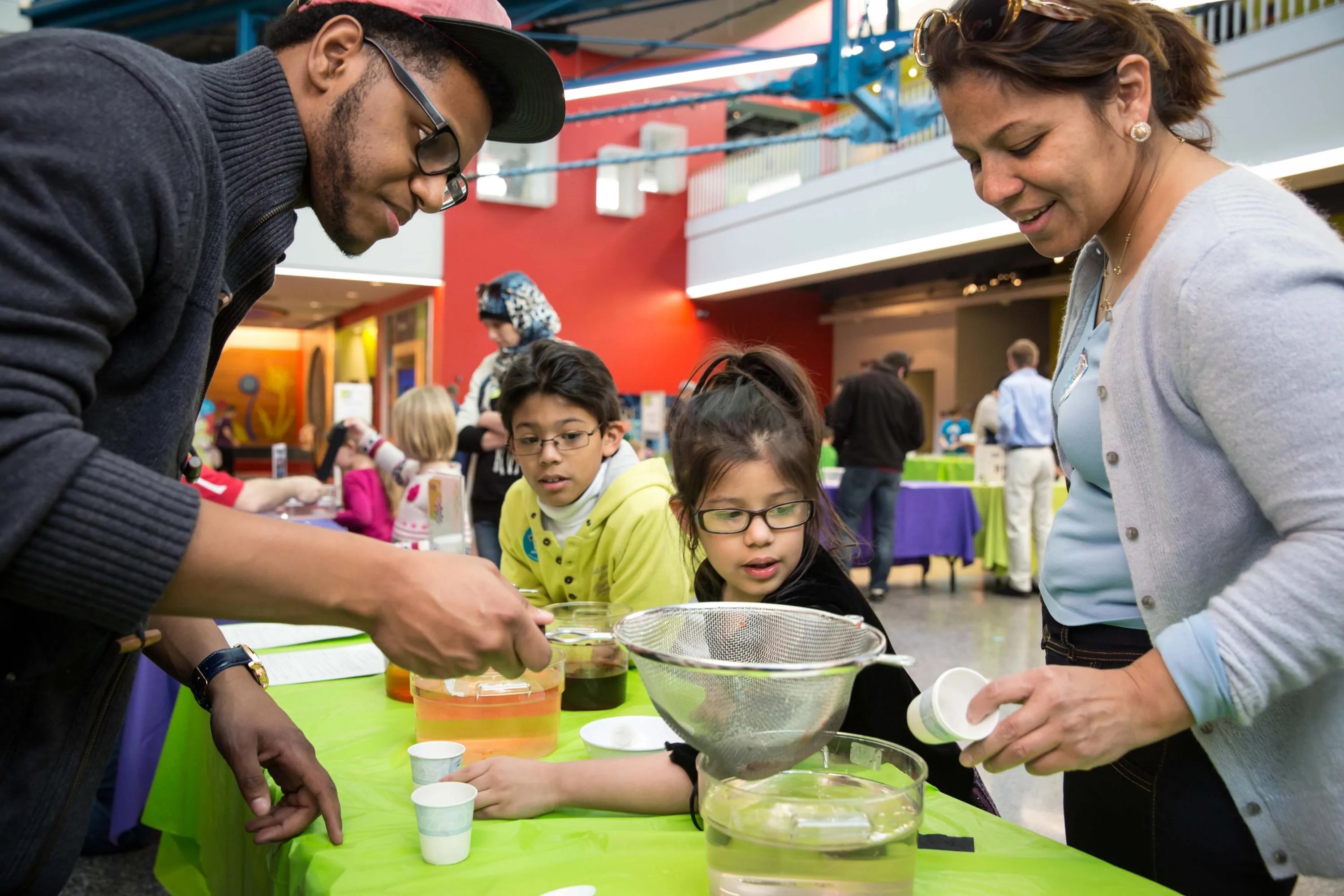 Photo of adults and kids at an activity table with a strainer and bowl of fluids