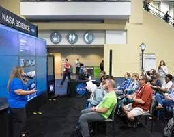 Infiniscope team member presenting Infiniscope learning experiences and teaching network on Hyperwall display within NASA booth. Audience seating filled with interested librarians.