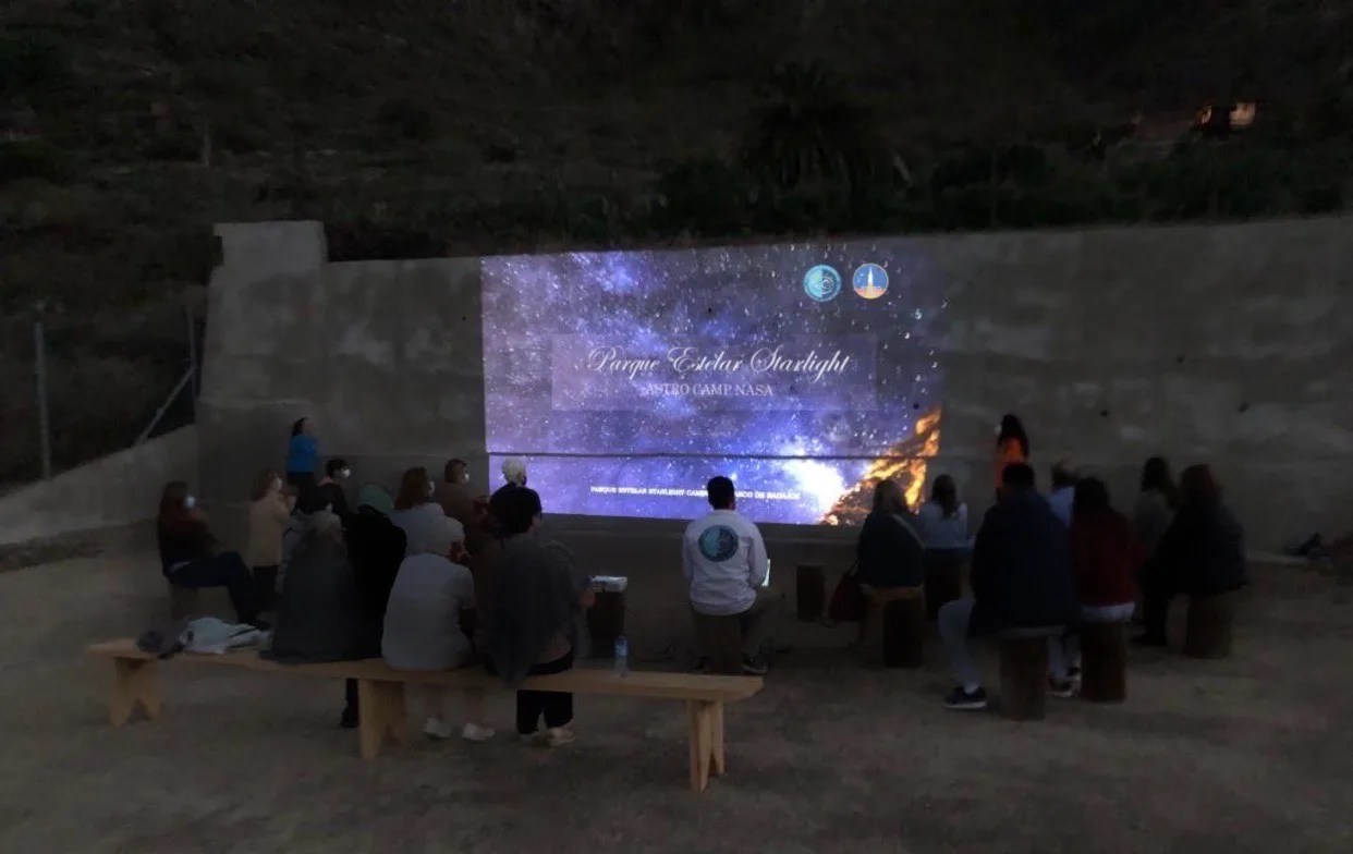 A dozen or so people on benches and cylindrical seats watch a presentation projected on a large concrete wall.
