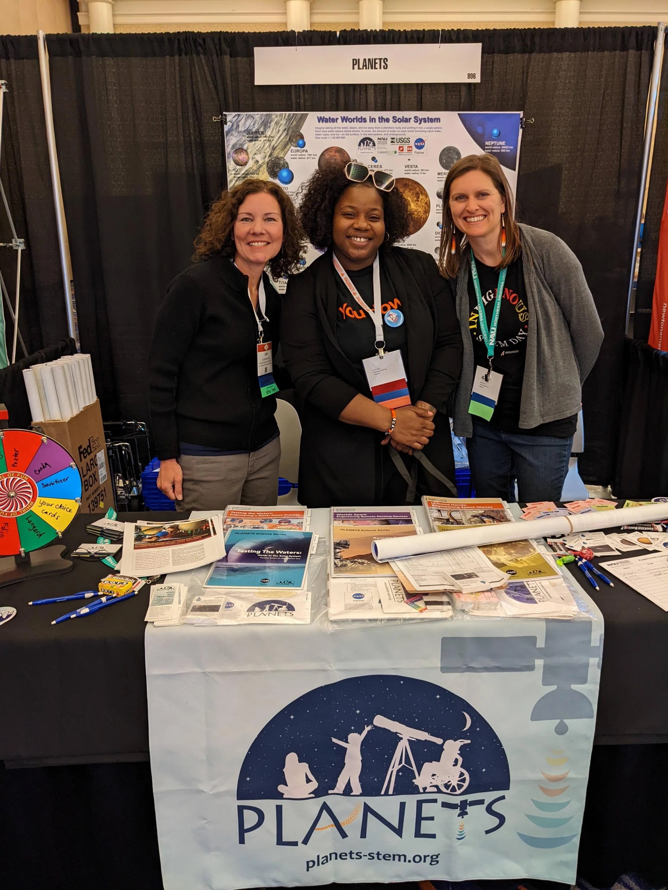 Three people standing behind an exhibit table draped with the PLANETS banner promoting the PLANETS program at the 2022 National Afterschool Association convention in Las Vegas.