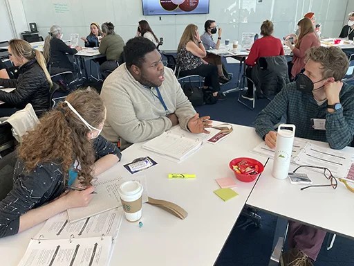 Three workshop participants are sitting at a table facing each other as they discuss the importance of forming authentic partnerships as one step in engaging diverse audiences. The participant in the middle of the image was speaking as the picture was taken.