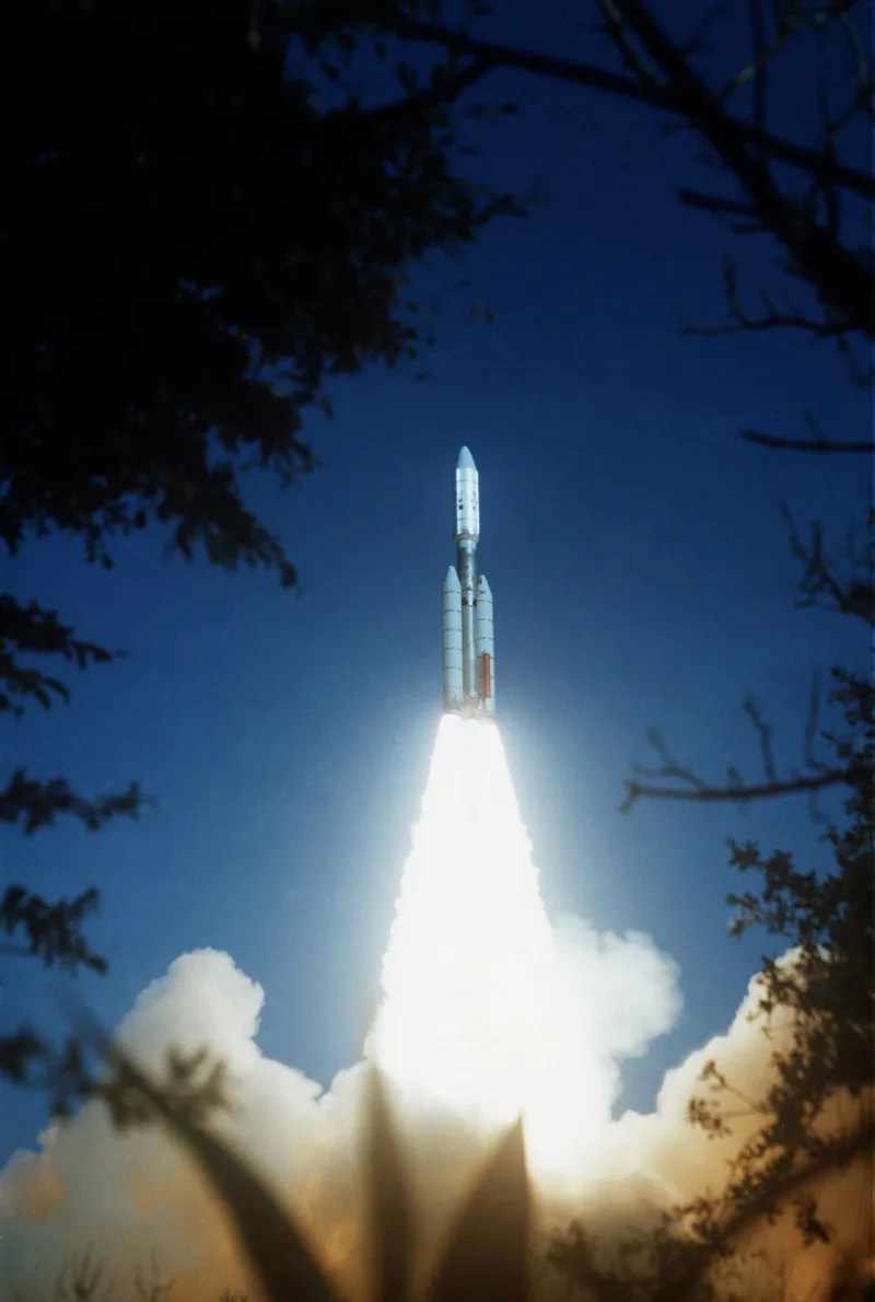 A large rocket carrying NASA's Voyager 2 spacecraft is seen through the trees as it lifts off with fire and smoke below it.