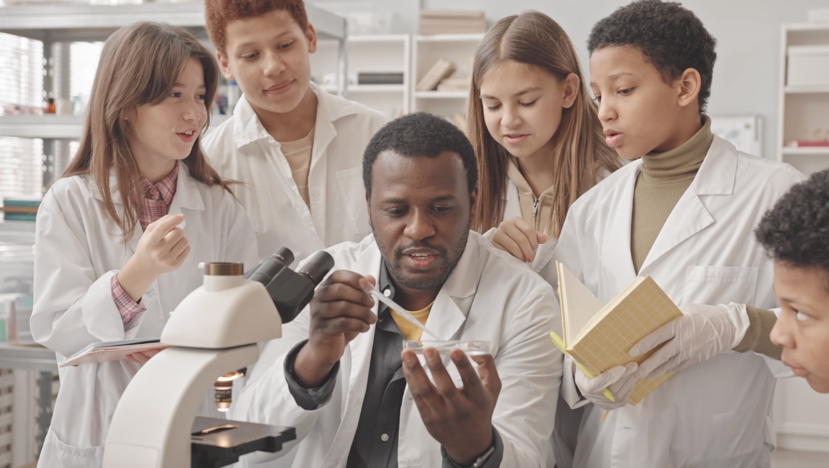 Screenshot from the video showing students as they gather around a scientist as he prepares a slide to be viewed under a microscope, which is part of studying how radiation affects humans in spaceflight.