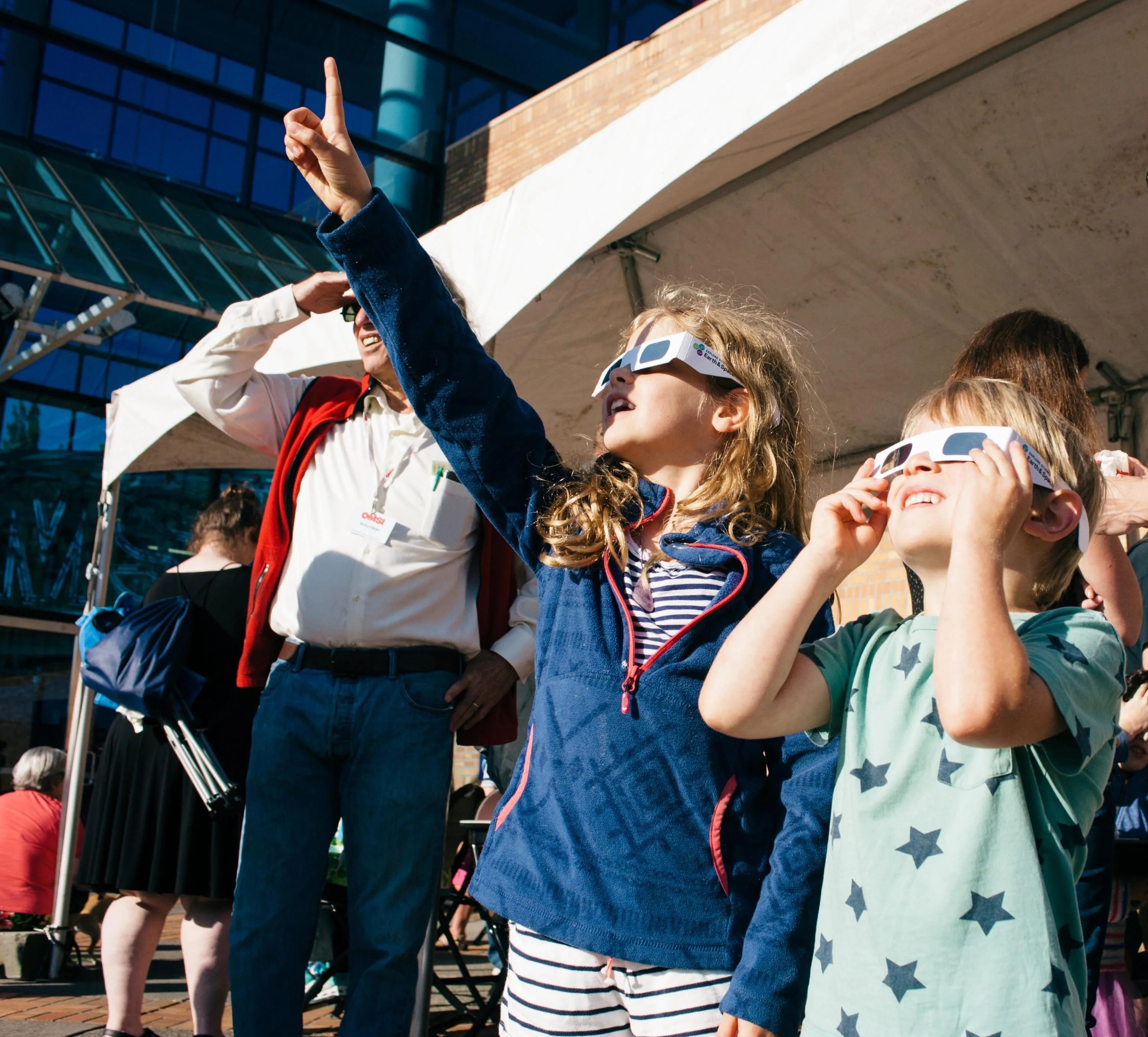 This photo shows families at a museum event, two young children in particular, safely viewing the 2017 solar eclipse while wearing safety glasses and pointing to the sky.