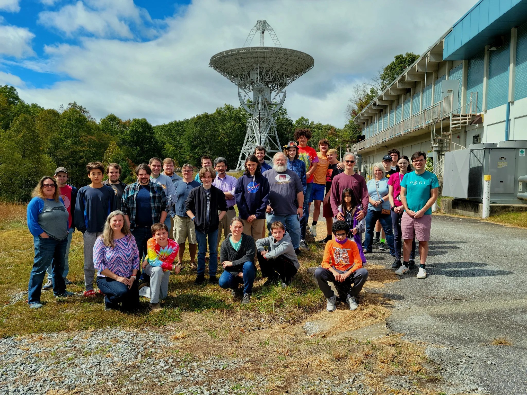 Photo of a group of 30 people posing for a photo in the grass in front of a large radio tower.