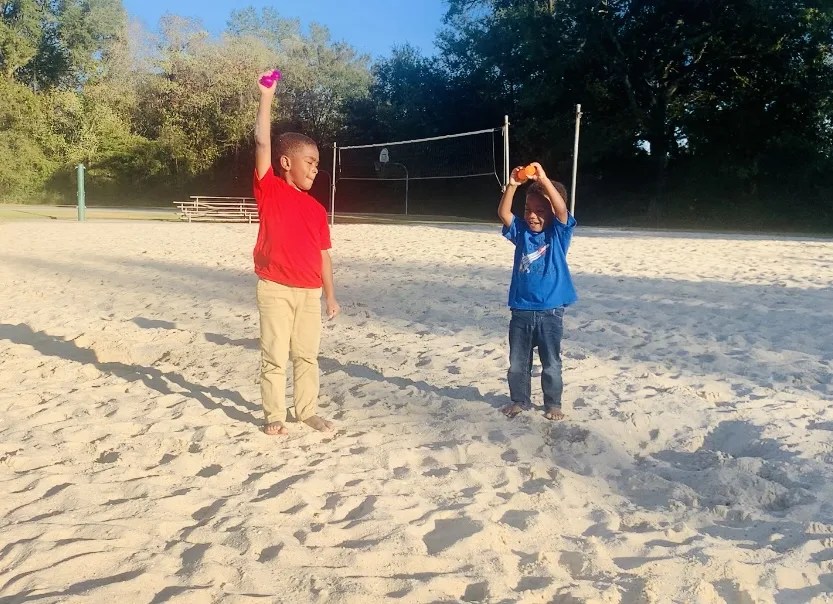 Two brothers stand on a sandy beach and hold brightly colored plastic eggs above their heads, ready to drop.