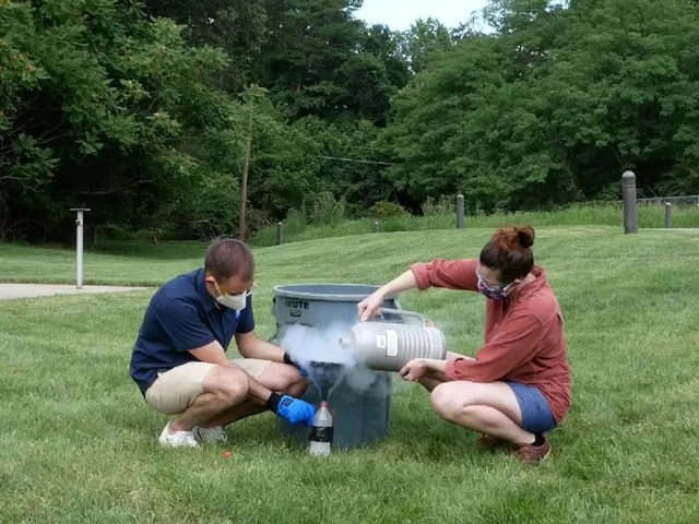 Two people squat down next to a large grey trash bin. The person on the right is pouring a gaseous substance into a funnel.