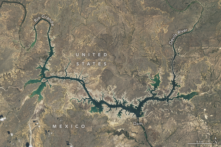 gray-brown landscape surrounds the Rio Grande and Devils rivers converging at a dam in the bottom right of the image and entering from the top left and top right of the image respectively. The dammed waterway is narrow and the banks, in many places are paler than the surrounding land.