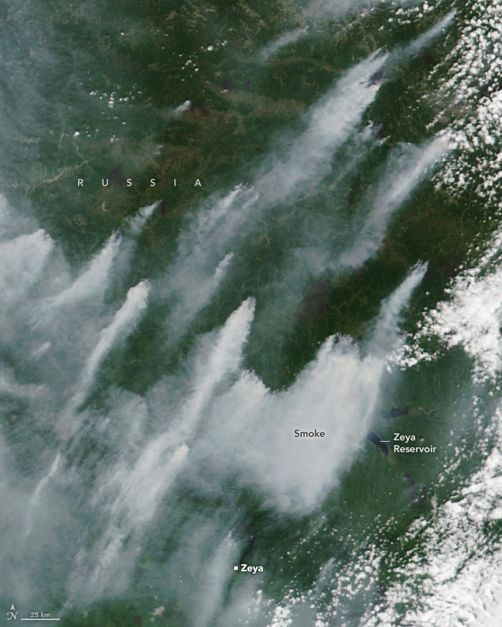 A satellite image of a very green landscape has numerous points of origin for smoke. The smoke is moving toward the bottom left of the image from these many points of origin and nearly block the land from view in the bottom left part of the image. Along the right edge are some scattered clouds, distinctly whiter than the blue-gray smoke.