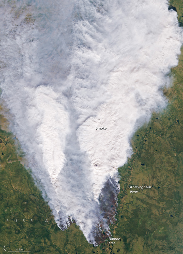 A large v shaped cloud of white smoke fills the upper part of the image and narrows down to the area of origin. At the origin region some land can be seen in a dark brown color compared to the surrounding landscape which is bright green.