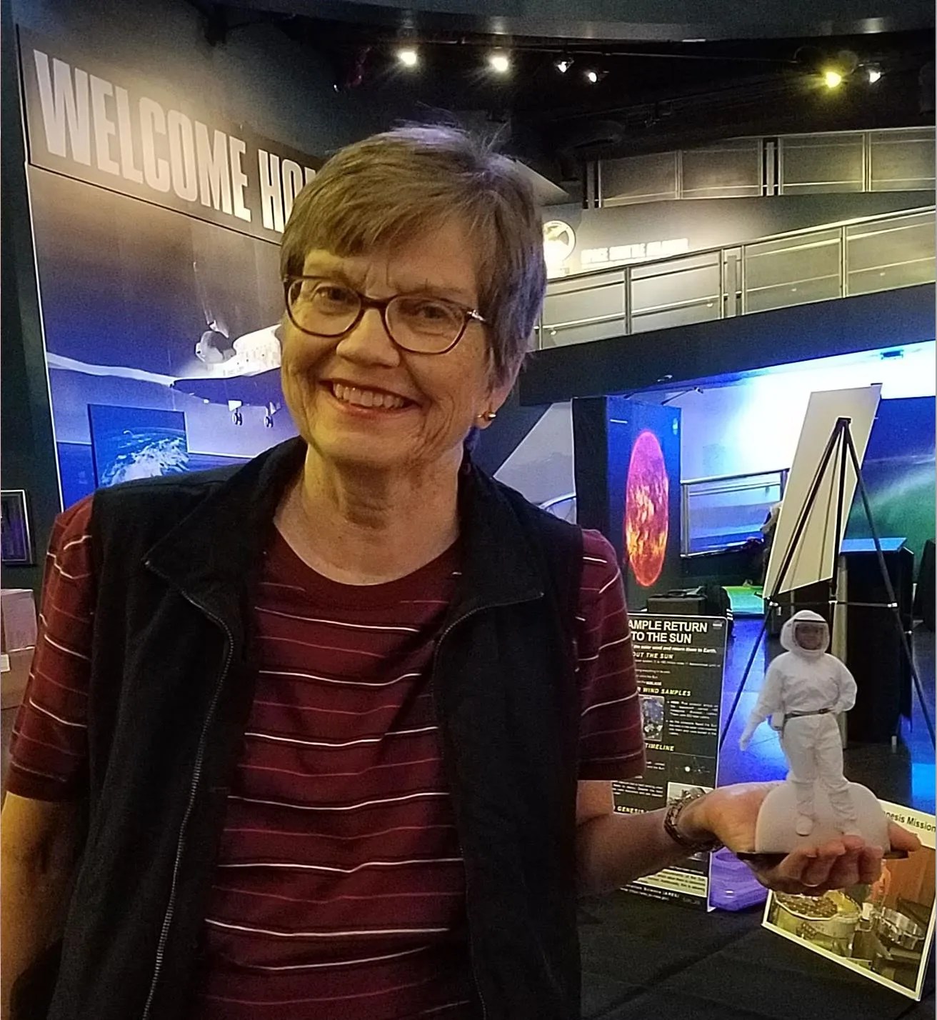 A woman with short hair and glasses holding a figure of a scientist in a white lab suit and helmet.