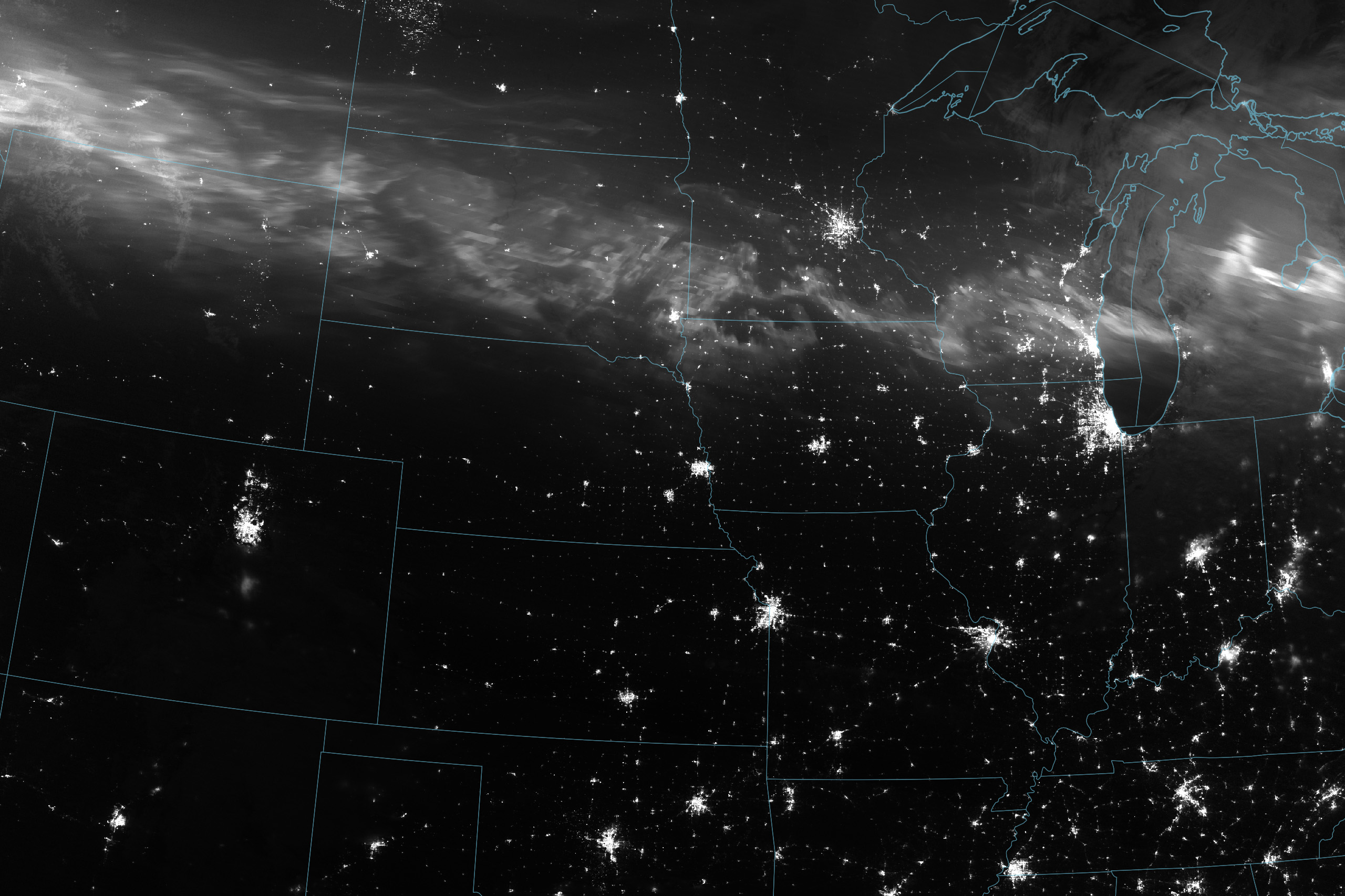 A nighttime satellite view of the United states, with state boundary overlays, centering the Iowa-Nebraska-Missourri border. The illumination of major city lights can be seen, most prominently Chicago in the upper middle right of the image. Just North of that you can see the densest part of a white haze that constitutes the Aurora can be seen. It spans the width of the image and is brightest on the far left over Montana. The belt of light curves steadily over the more northern US states with swirls of higher density.