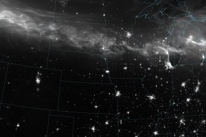 A nighttime satellite view of the United states, with state boundary overlays, centering the Iowa-Nebraska-Missourri border. The illumination of major city lights can be seen, most prominently Chicago in the upper middle right of the image. Just North of that you can see the densest part of a white haze that constitutes the Aurora can be seen. It spans the width of the image and is brightest on the far left over Montana. The belt of light curves steadily over the more northern US states with swirls of higher density.