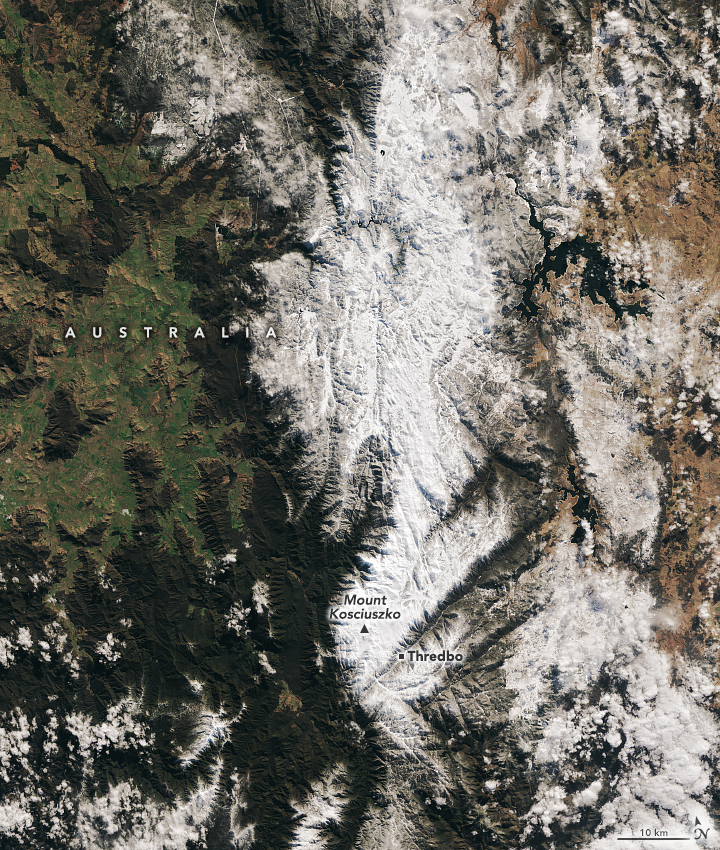 Mountain peaks run vertically down this image, covered in snow. To the left of the snowy mountains are dark green peaks and light green valleys. To the right of the snowy peaks the land appears more brown through the sporadic cloud coverage.