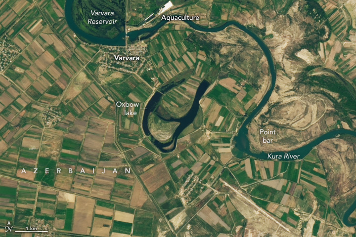 The image is primarily partitioned crop fields. In the center of the image is an Oxbow lake, and the Kura River meanders along the top of the image and down to the right. In the top center left the Varvara Reservoir, darkens the foliage above the river. On the opposite side of the river is the town of Varvara. An aquaculture facility is fed by and sits to the right of the Varvara Reservoir. On the right side of the image a point bar is created where the river meanders away from the side of the land.