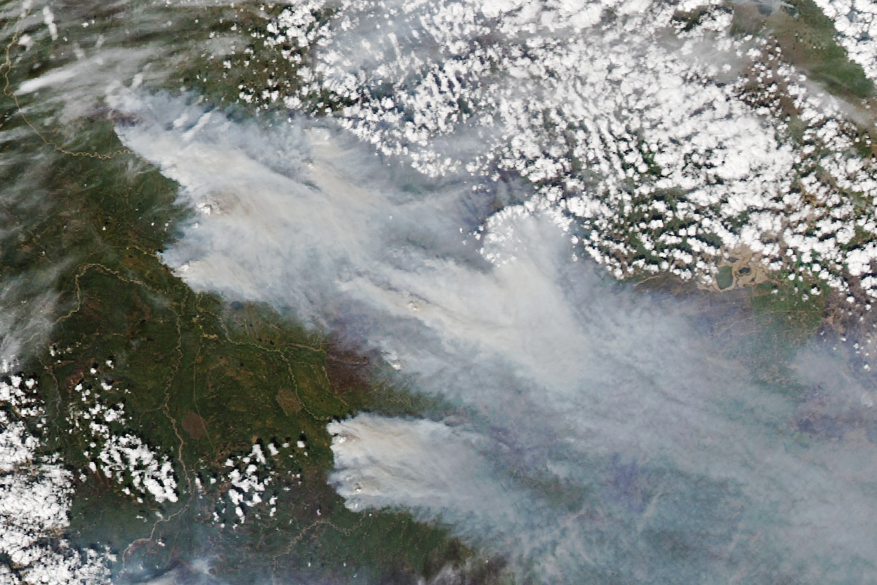 Satellite photo of a deep green landscape. The top right corner is mostly covered by a patchwork of small altocumulus clouds, the land showing between not significant enough to make any discernable insights. A much smaller patch of similar clouds are also found in the lower left. Streaked between the two patches of clouds is a significant amount of smoke, discernable origin points near the top left, mid center left where Fort Nelson is, and lower center. The smoke is blown to the right of the image and the origin points are opaque, but the bottom right appears somewhat translucent and imparts a somewhat blue hue to the ground seen below.