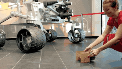 Animation of a woman spinning the wheels of a cardboard rover, with a larger model rover in the background.