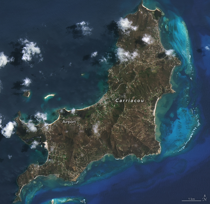 Amid coastal turquoise blue waters a remarkably brown colored island exists in two connected segments, one vertical, the tip at the top right of the image, and then branching left at nearly 90 degrees is another relatively narrow segment of land. The full island is in view and individual roadways remain visible in this satellite image.