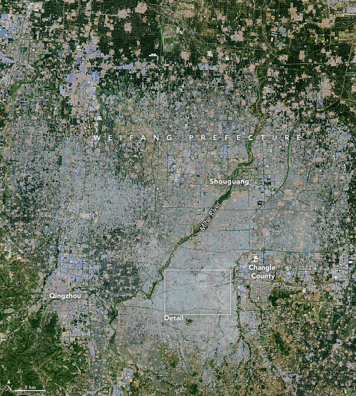 Land centering around a streak of green that make up the banks of the Mihe River, which runs between the top center right of the image to the bottom center left of the image, is almost entirely gray, the color of a greenhouse from satellite images. on the outermost perimeters of the image there are smatterings of green, but some small pockets of greenhouses as well.