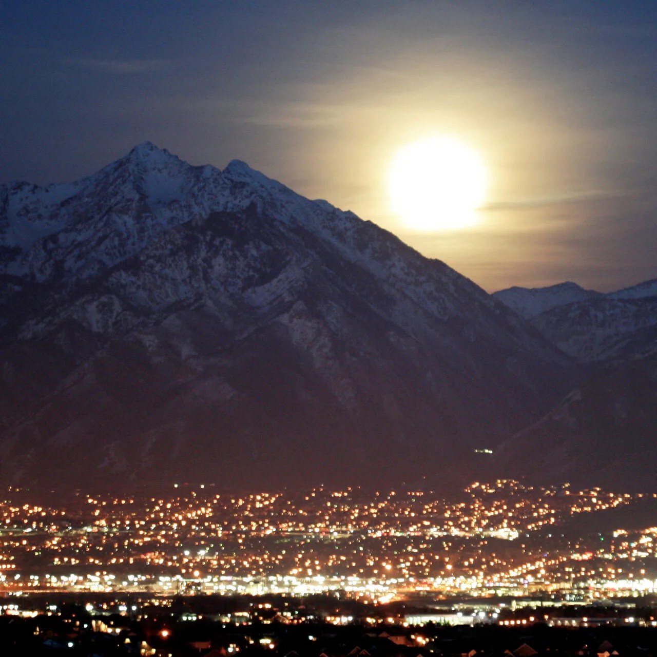 Bright moon rising over mountains with city lights in the foreground.
