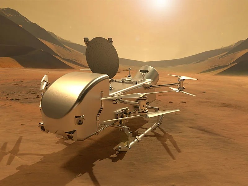 A shiny spacecraft sits on a red-brown sandy surface. Sand dunes are shown rising high in the distance.