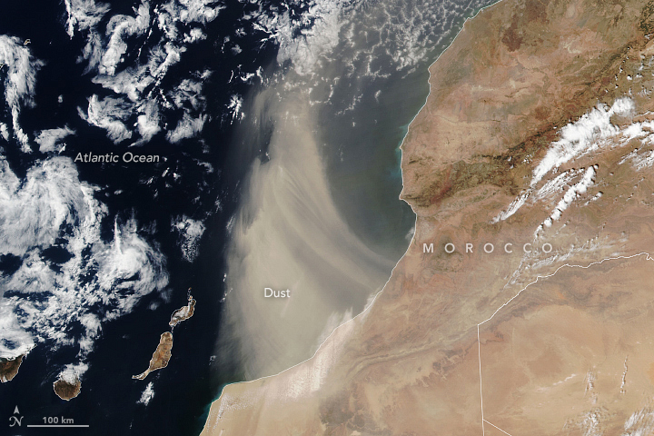 The Moroccan coastline runs diagonally across the image. The land is on the right and largely beige and brown in color. The Atlantic Ocean on the left along with some small islands. Significant cloud coverage is interspersed over the dark blue ocean waters. Light colored dust blows off the land in the center of the image, obscuring the ocean below somewhat.