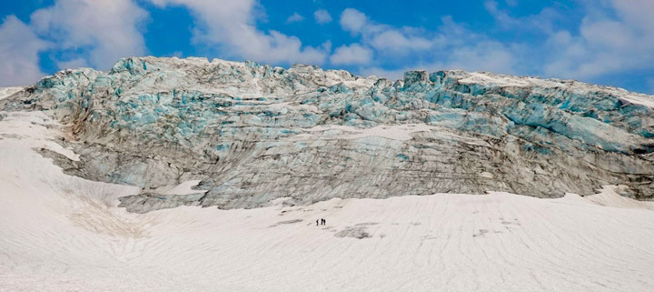 Image of a massive glacier dwarfing three people at its edge of folded ice surrounded by snow.
