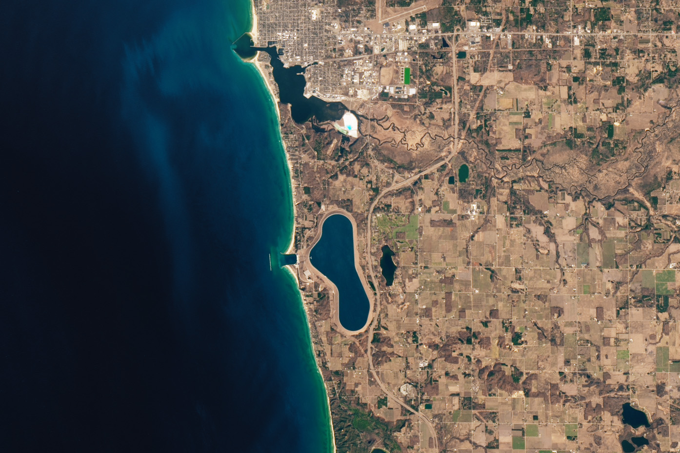 Lake Michigan is shown left in a rich blue and the state of Michigan is shown left. The land appears to largely be farmland in gridded fields, but most notably just barely inland from the coasts is an oblong lake stretched top to bottom parallel to lake Michigan. The two lakes are joined by a dam. The Natural coastline of Michigan is disturbed by a carveout for the dam outflow with a thin line of the breakwater visible in the lake directly in front of the dam