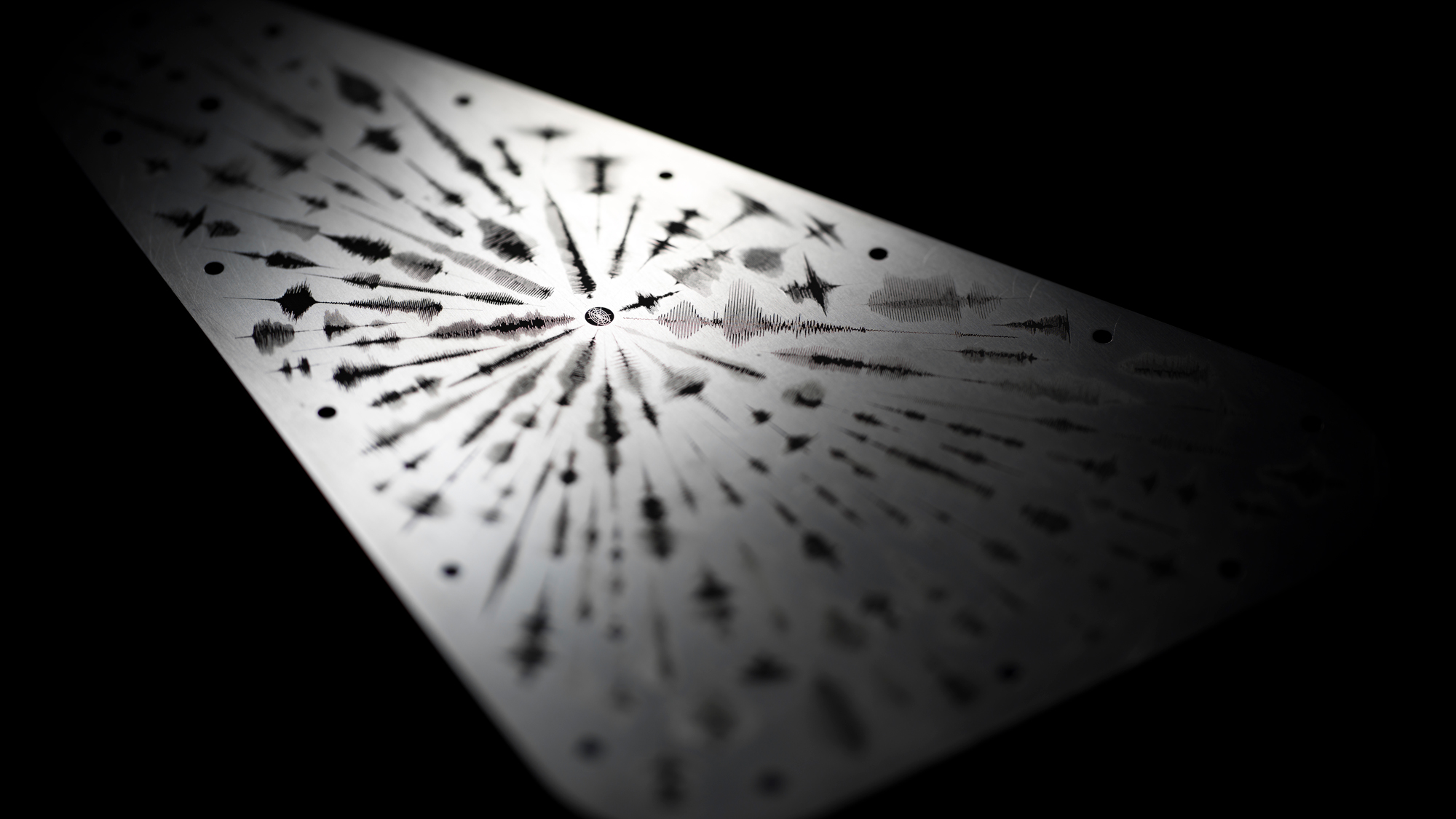 An image of the Europa Clipper vault plate, highlighted from the upper left corner casting shadows that emphasize engraved waveforms emanating from a central point symbolizing the word 'water' in multiple languages. The waveforms present as audio frequency waves. The vault plate is metal silver and the engravings are black, set against a black background.
