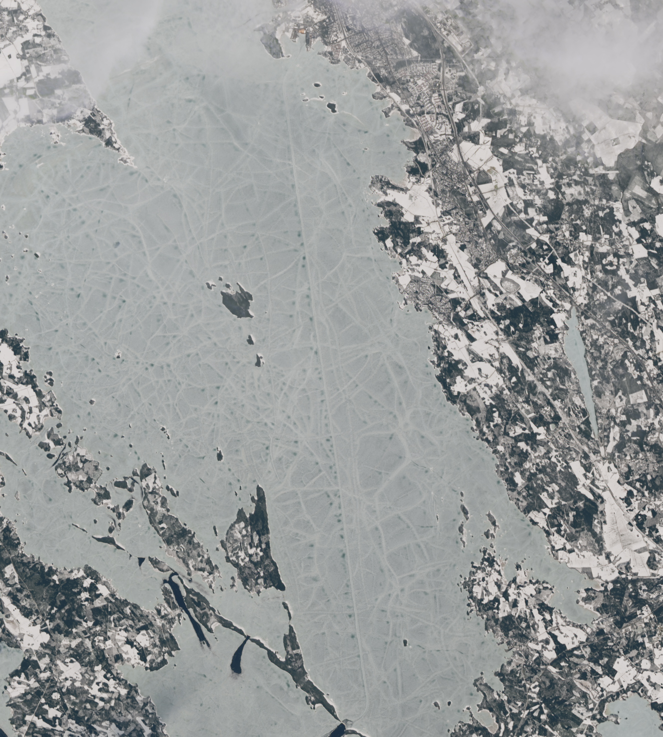 An icy lake centers the image with mainland to the right and sporadic islands and some smaller inhabited peninsulas to the left. The land is covered in snow. A vertical line of bright white ice is seen south of the lone visible population center.
