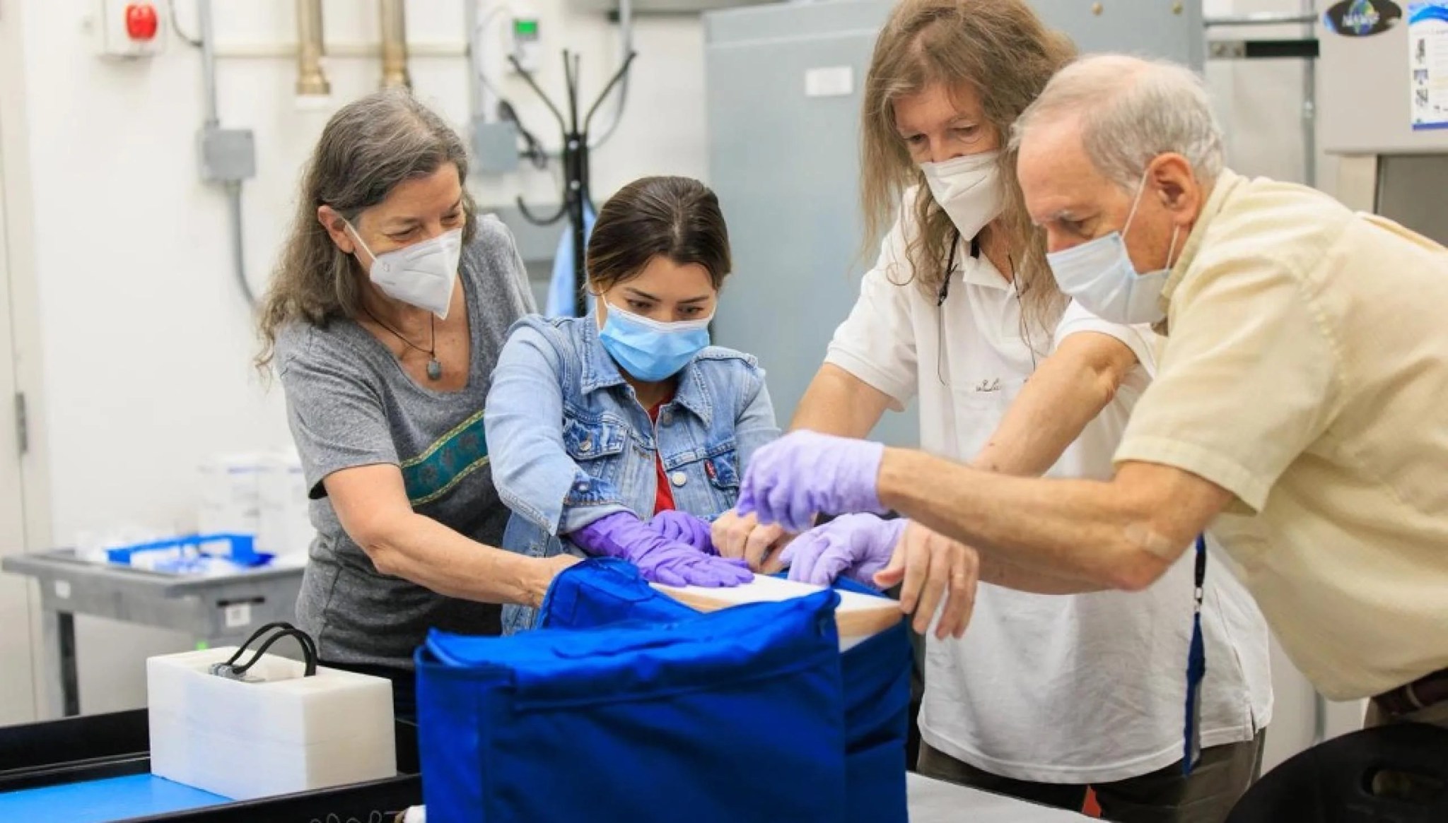 Three women and one man are huddled around a box. They are wearing masks and gloves and are removing a medium-sized rectangular box from a cerulean blue canvas bag.