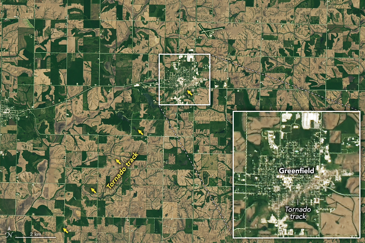 Satellite image of a town centers the image surrounded by gridded roads and farmland in dark green and brown. Diagonally from the bottom left into the town a faint discoloration of the landscape is visible which becomes pronounced when it intersects the town where the green yards and fields are discolored significantly with light brown dust and debris from the destruction. The town is zoomed in on via in a square superimposed over the lower right of the image.