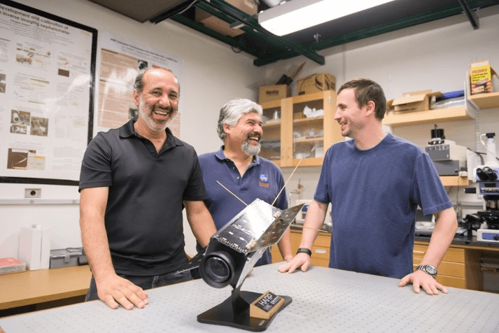 Photo of three men with smiles on their faces standing in a lab at a table with a full size model of HARP, a black rectangular boxy object on a pedestal