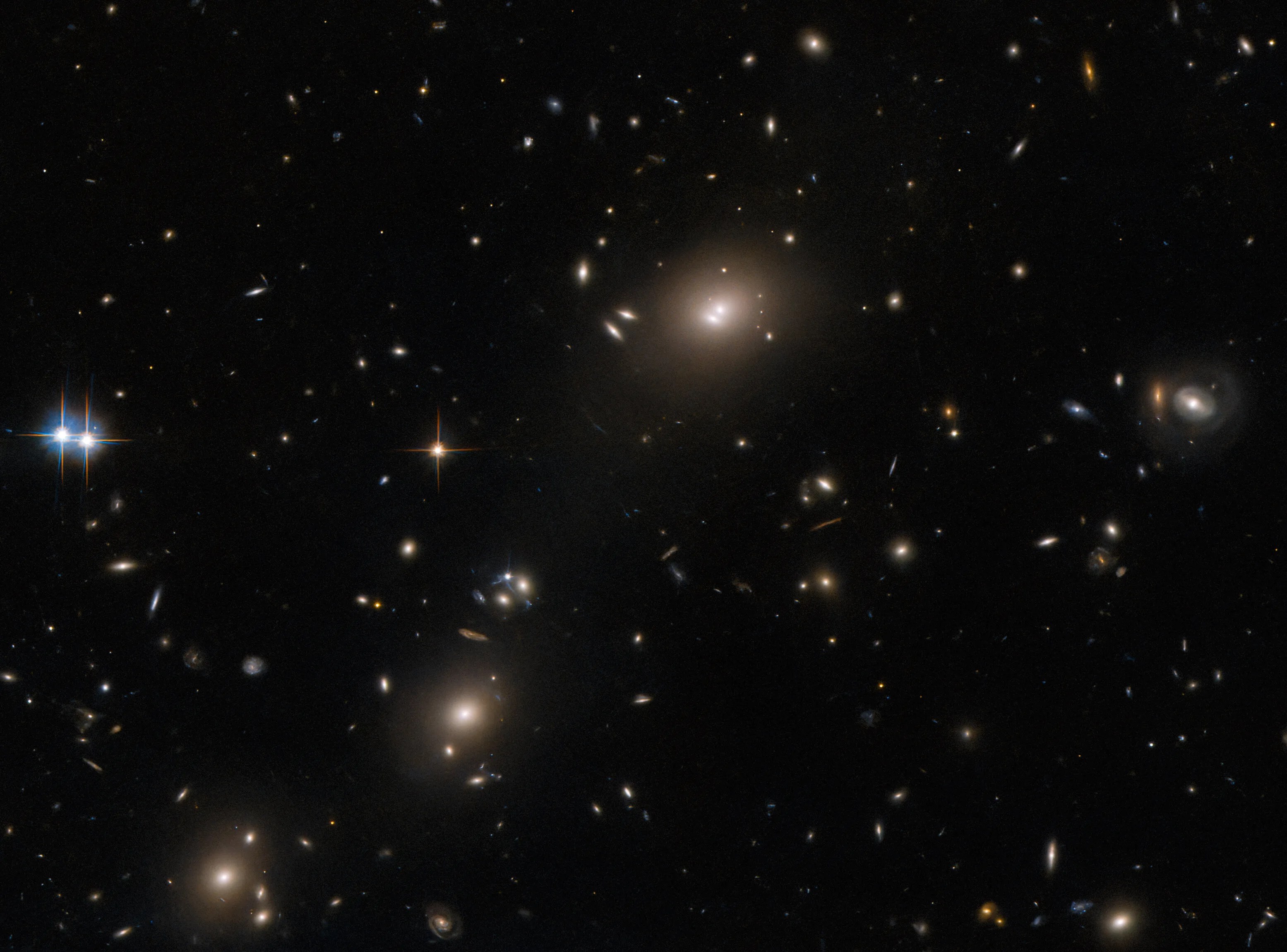 A collection of oval-shaped, elliptical galaxies. The largest has two neighboring bright spots in the core. It and two others look like galaxy clusters, with surrounding smaller galaxies. On the left edge of the image are two bright stars with four long spikes, and on the right edge is a small ring-shaped galaxy. Smaller stars and galaxies are spread evenly across the dark background.