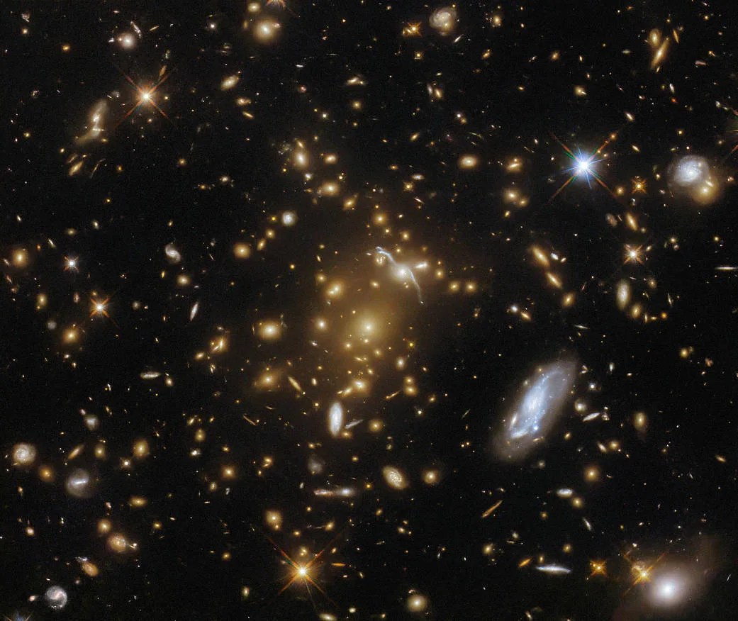 A cluster of large galaxies, surrounded by various stars and smaller galaxies on a dark background. The central cluster is mostly made of bright elliptical galaxies that are surrounded by a warm glow. Close to the cluster core is the stretched, distorted arc of a galaxy, gravitationally lensed by the cluster.