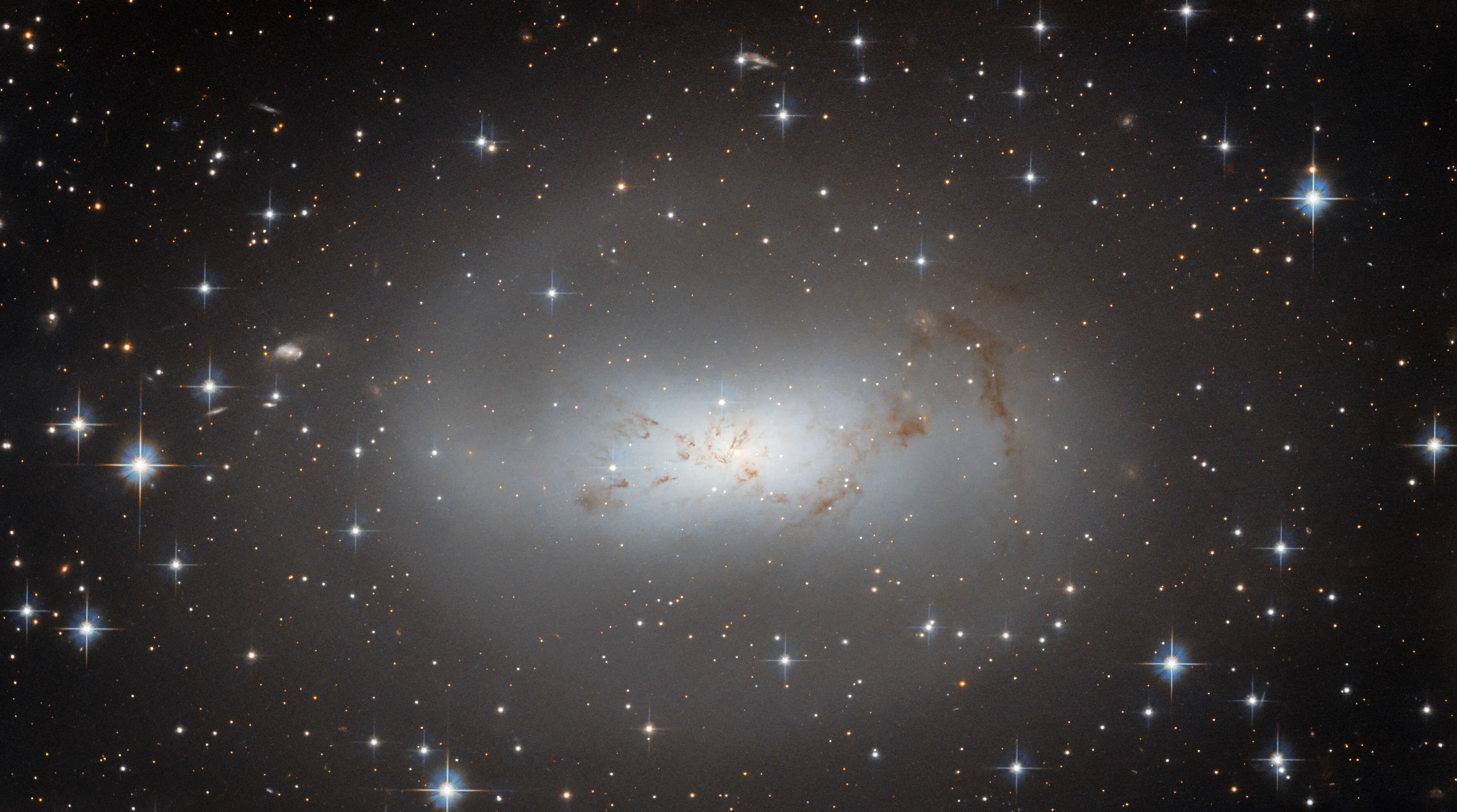 A galaxy, large and occupying most of the view from the center. The whole galaxy is made of smooth, diffuse light. In the center it is brighter and bluer, fading to a pale gray halo that is faint and see-through. The light forms an arm on one side that curls around the top. A couple threads of dark dust cross the center. Many stars shine around the galaxy, on a black background.