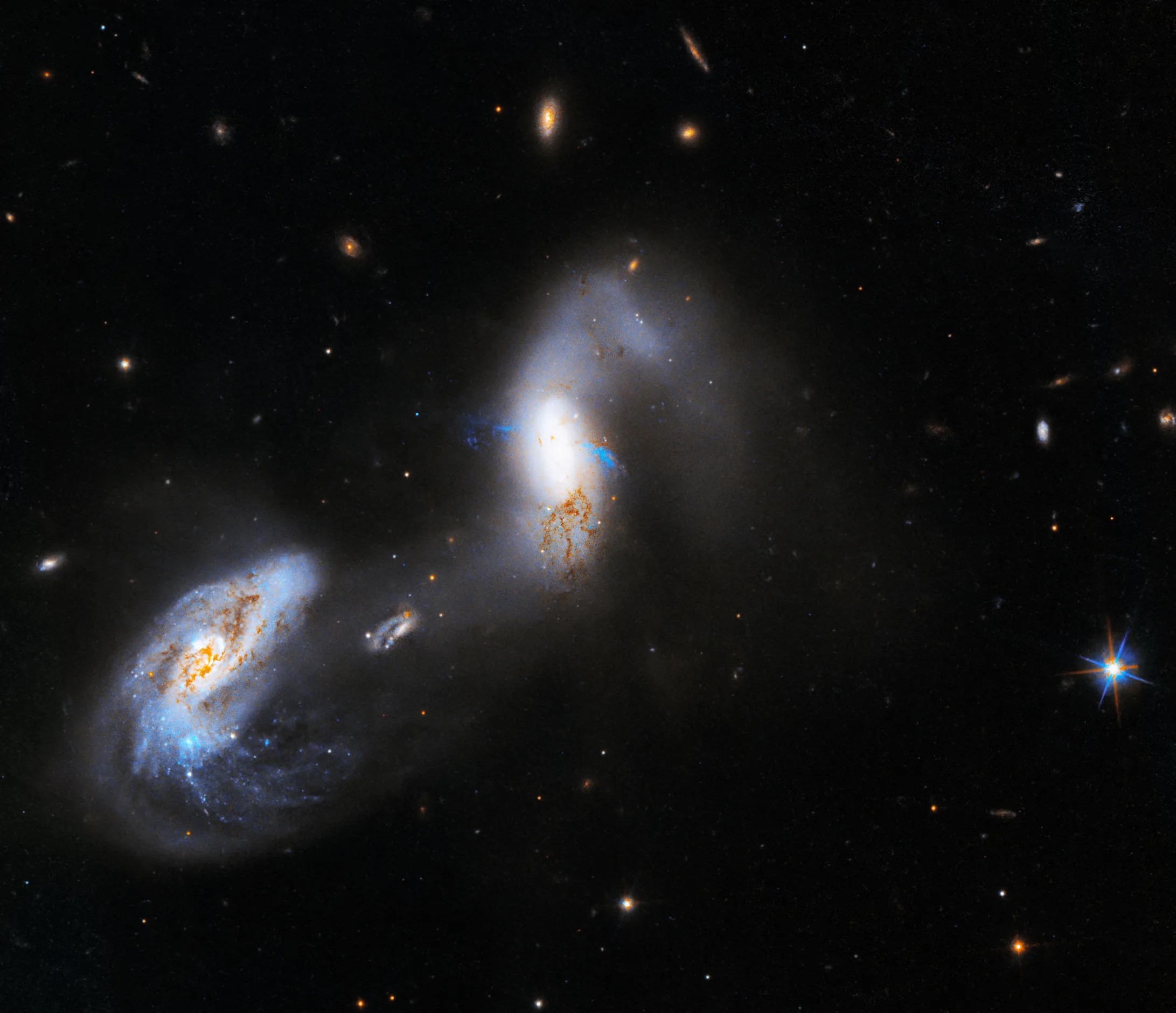 Two blue-white, comma-shaped galaxies: at upper-right and lower-left of center. Streams of diffuse gas stretch between them. Areas of reddish-brown dust dot galaxy at left, less so on galaxy at right. Black background dotted with stars, distant galaxies.