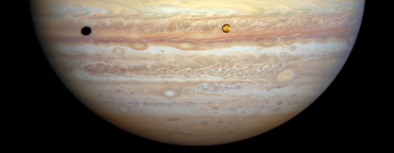 Lower half of Jupiter. Cloud bands of rusty-orange, dull yellow, white, and brown. Swirling ovals and turbulent waves of clouds bound by the bands. Yellow-orange moon, Io, just right of center. It casts its shadow toward the planet's left limb.