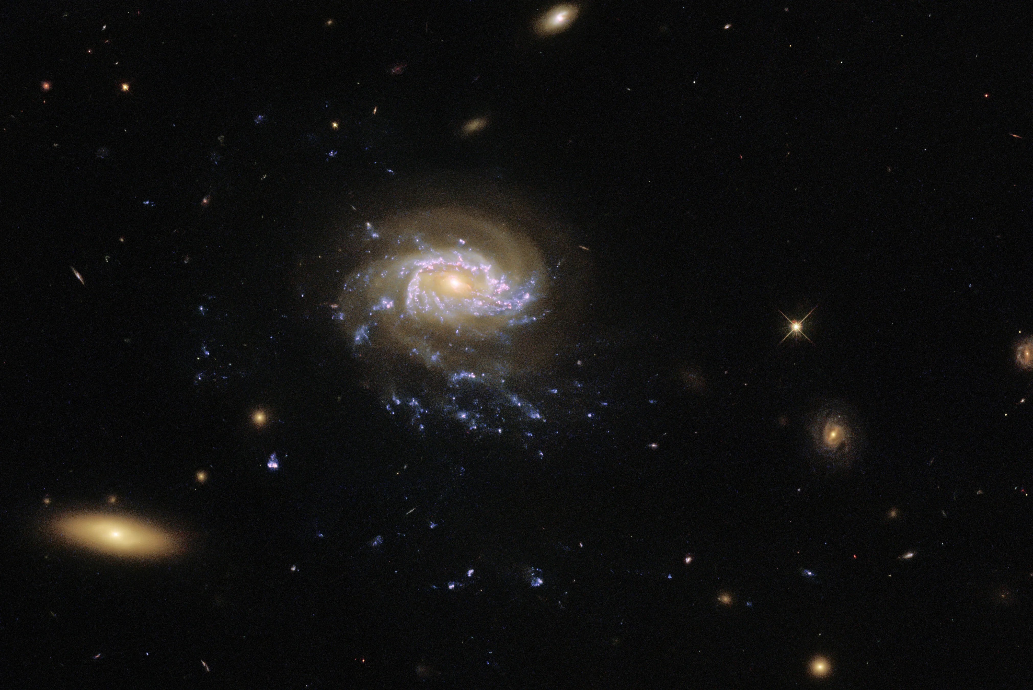 A spiral galaxy lies just off-centre. It has large, faint, reddish spiral arms and a bright, reddish core. These lie over two brighter blue spiral arms. These are patchy, with blotches of star formation. Long trails of these bright blotches trail down from the lower spiral arm, resembling tendrils. The background is black, lightly scattered with small galaxies and stars, and a larger elliptical galaxy in one corner.