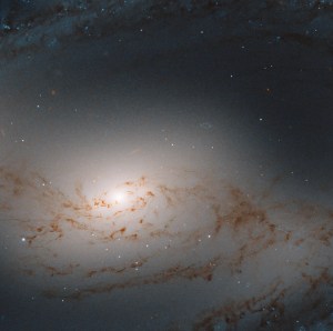 Haze of light. Bright core of the galaxy just left of center. A smattering of rusty-brown dust clouds in the foreground.