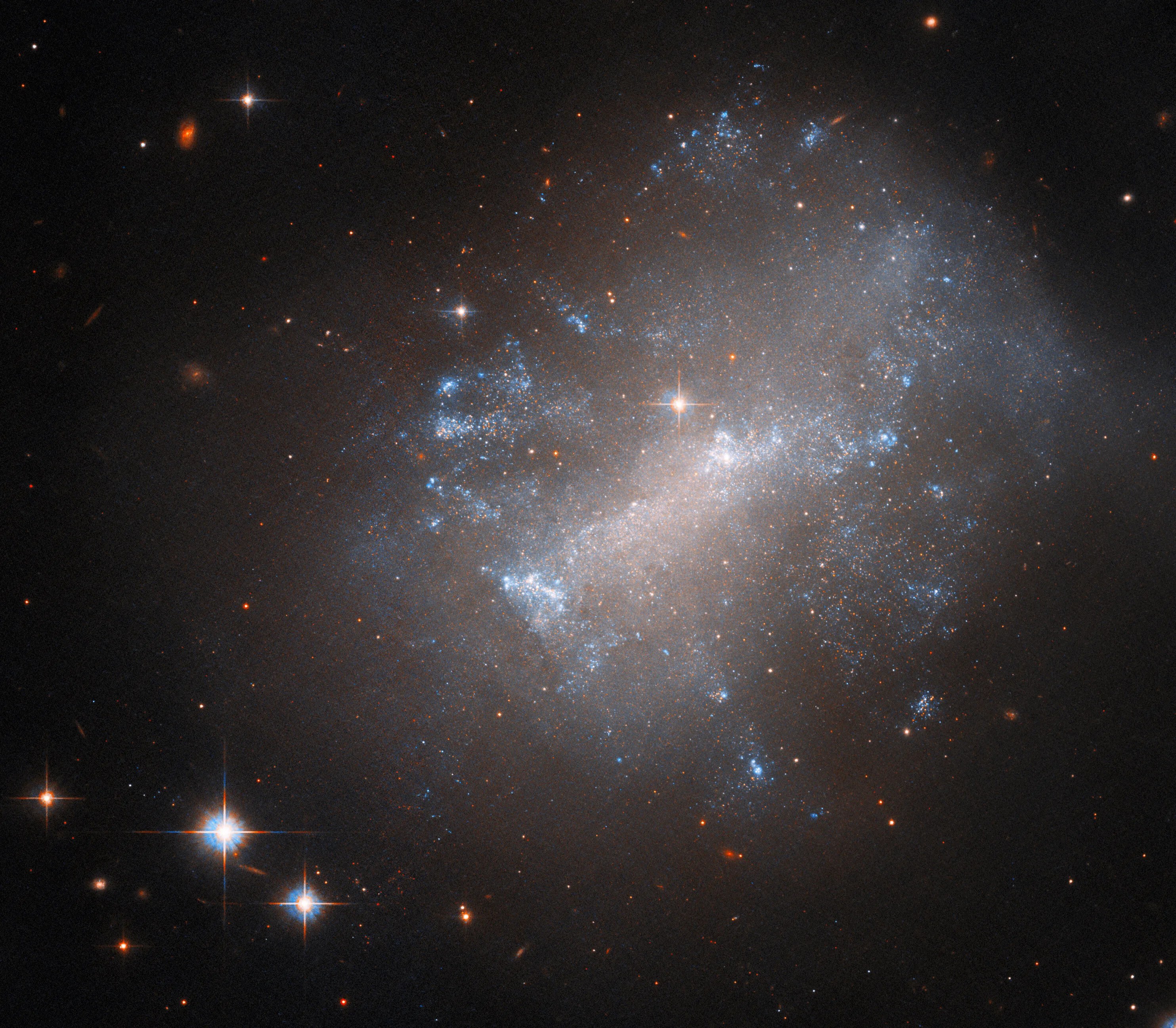 A galaxy fills up most of the frame from the right. It is fuzzy and diffuse but made up of numerous tiny stars. In the core, the stars merge into a glowing bar shape. The gas and stars in the galaxy vary between warm and cool colors. They are spread over a large area, the colors mixing like clouds. The glow of the galaxy fades into a black background, with a few stars and small, distant galaxies.