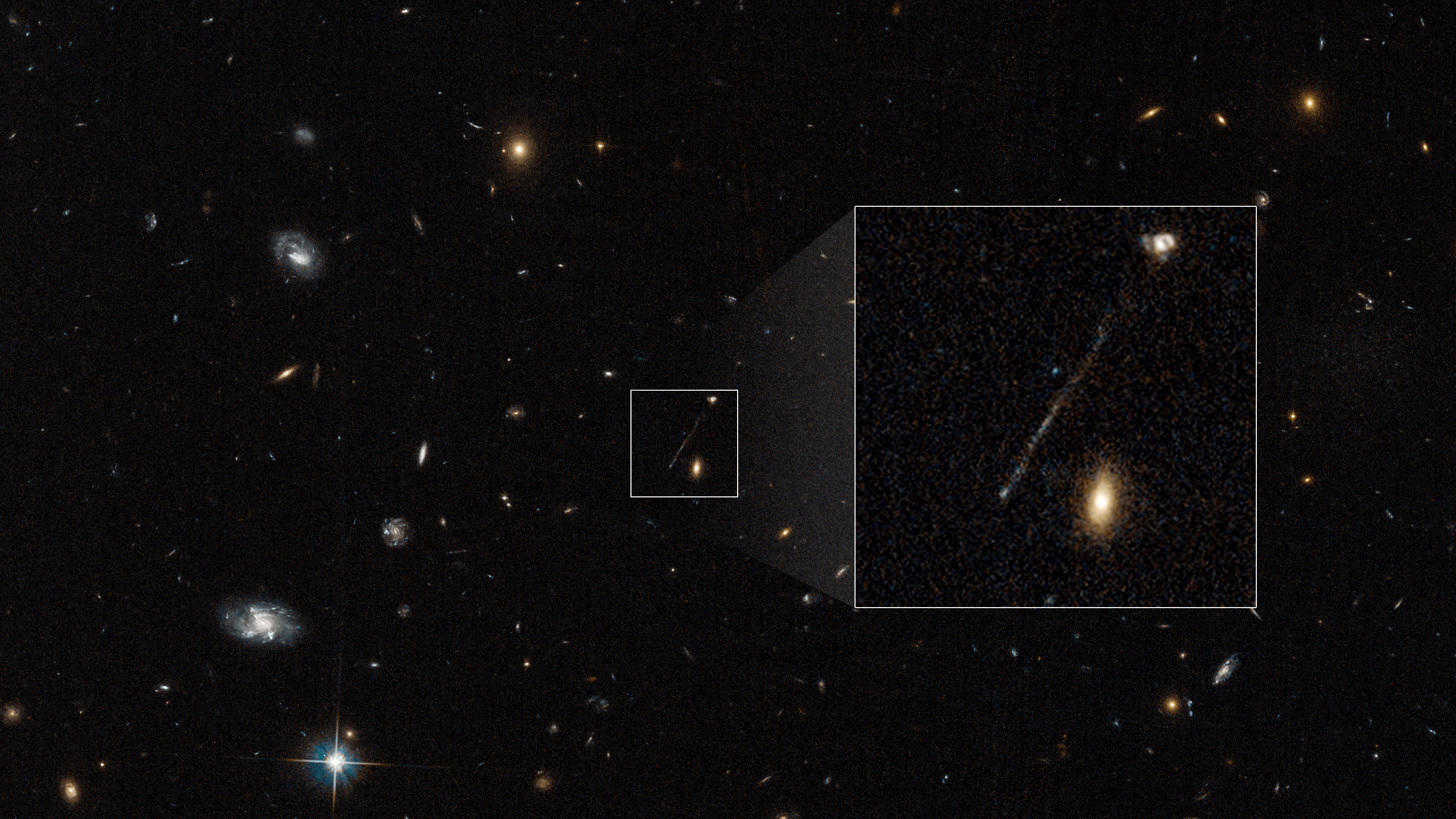 Hubble image of black, deep-space field with white, yellow, and reddish galaxies. Image center: small, white-bordered, boxed area that contains one, long, thin, diagonal streak of whitish-blue stars. Two galaxies also reside within the box.