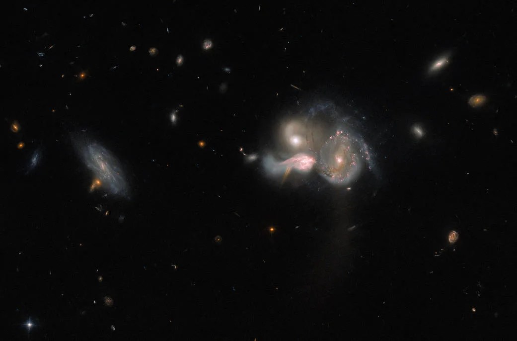 Three galaxies stand together just right of centre. They are close enough that they appear to be merging into one. Their shapes are distorted, with strands of gas and dust running between them. Each is emitting a lot of light. Further to the left is an unconnected, dimmer spiral galaxy. The background is dark, with a few smaller, dim and faint galaxies and a couple of stars.