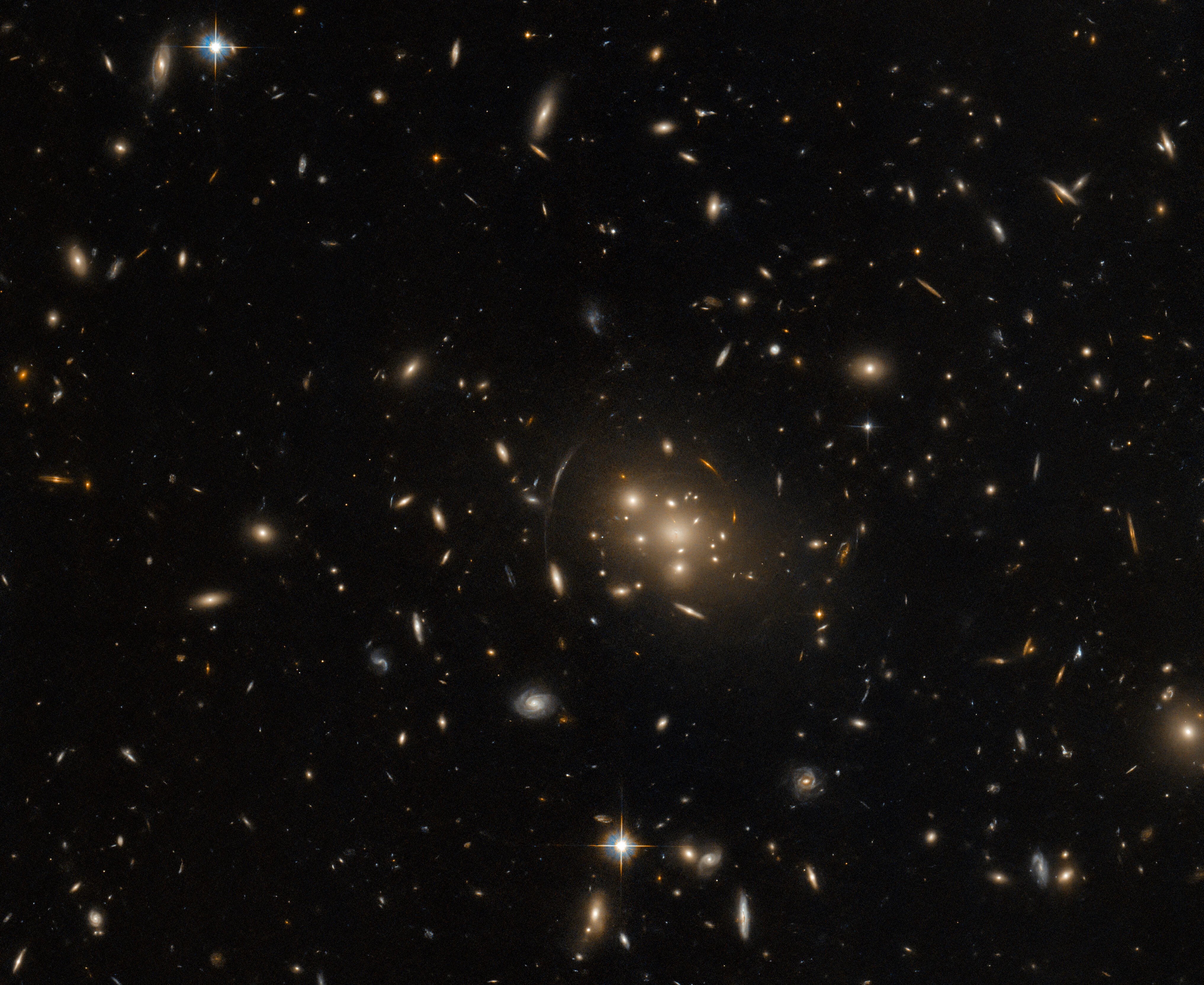 A cluster of large galaxies, surrounded by various stars and smaller galaxies on a dark background. The central cluster is mostly made of bright elliptical galaxies that are surrounded by a warm glow. Nearby the cluster is the stretched, distorted arc of a galaxy, gravitationally lensed by the cluster.