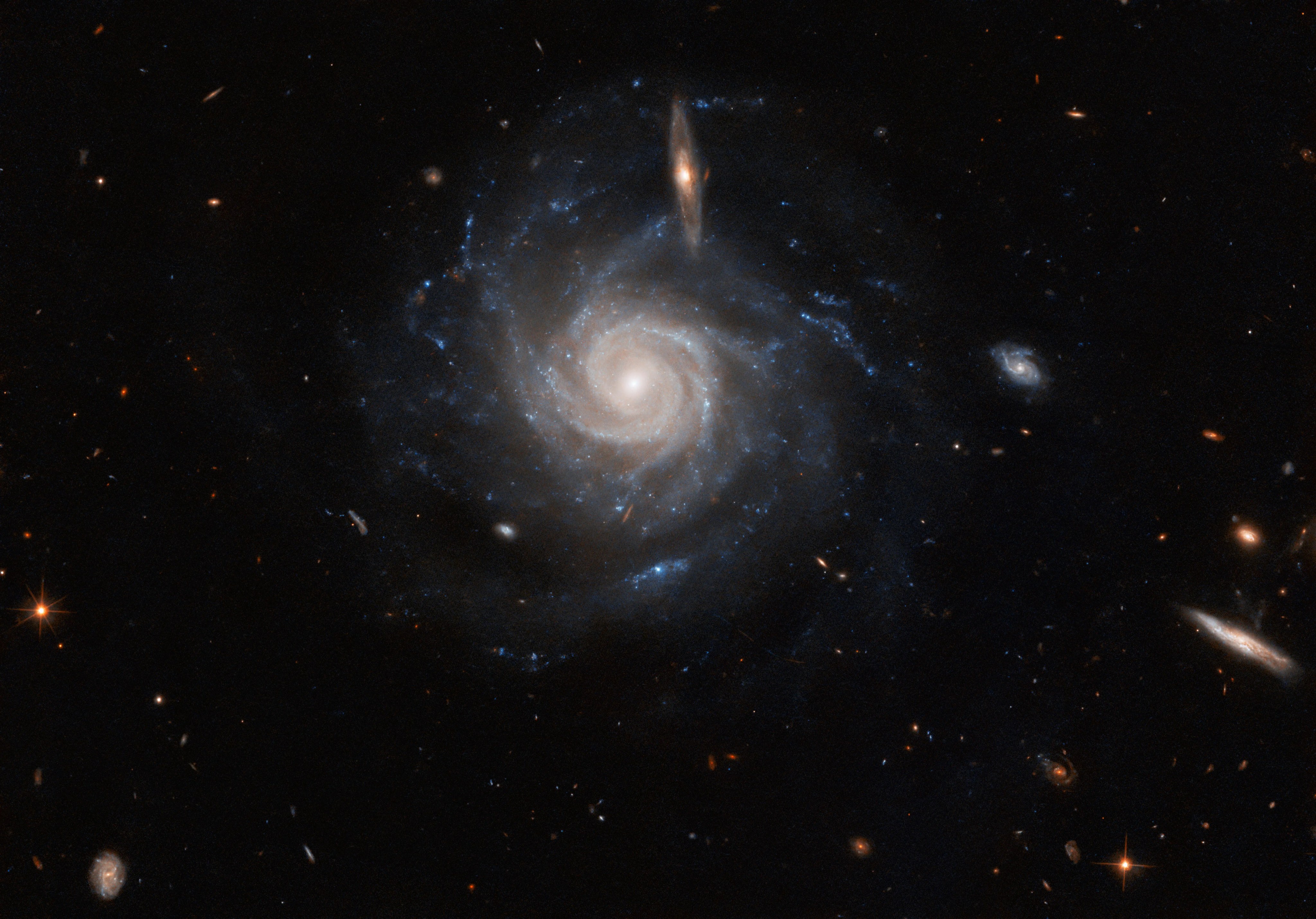A large spiral galaxy. It has many narrow arms that are tightly-twisted in the center, but at the ends they point out in different directions. The galaxy’s core glows brightly, while its disc is mostly faint, but with bright blue spots throughout the arms. A few smaller spiral galaxies at varying angles are visible in front, and it is surrounded by other tiny stars and galaxies, on a black background.